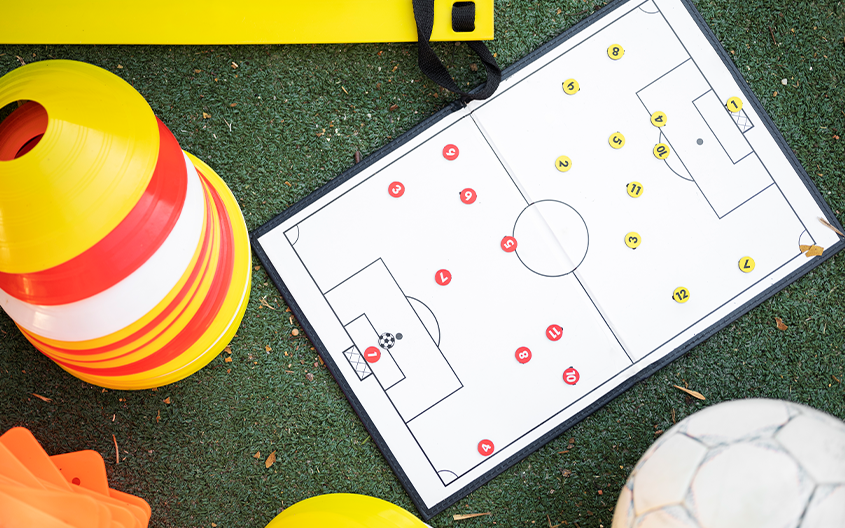 An overhead shot of a football tactical coaching board lying on green turf, displaying a pitch diagram arranged with red and yellow numbered player markers. Surrounding the board are training tools including a stack of yellow and orange cones, a yellow agility ladder rung, and a football.
