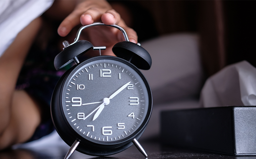 A close-up shot of a black twin-bell vintage alarm clock sitting on a nightstand, with a person's hand reaching from bed to turn it off, illustrating sleep habits for athlete recovery.