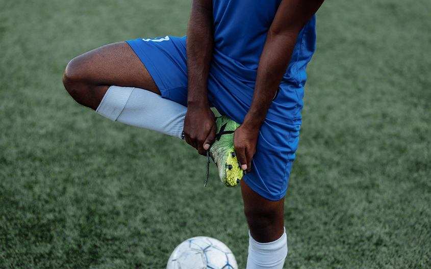 A football player wearing a blue kit standing on an artificial grass pitch, holding their ankle and stretching their leg to prevent injury, with a football at their feet.