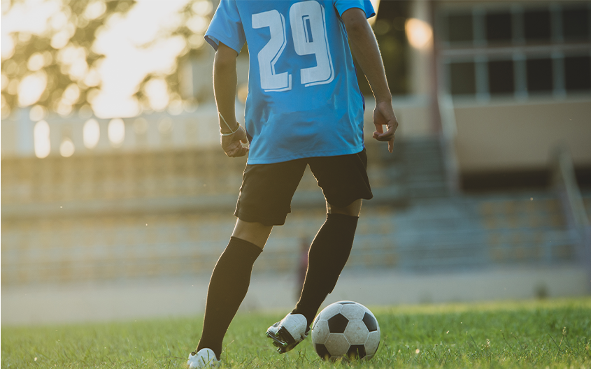 A low-angle action shot from behind a football player wearing a light blue number 29 jersey and black shorts, using his cleats to control a football on a green grass field.